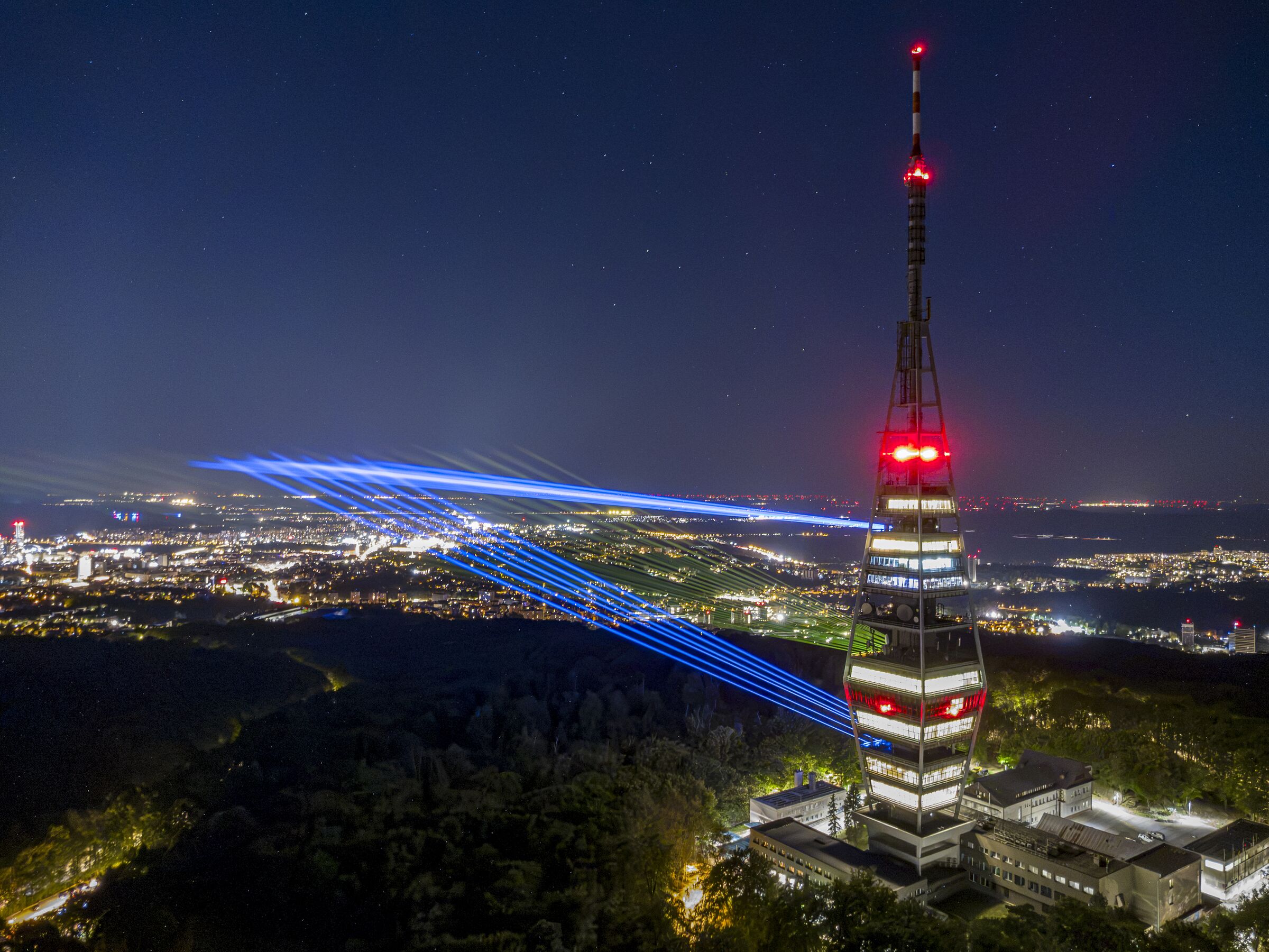 Night over Bratislava from 120 metres: blue laser beams fan out from the lit Kamzík TV tower across the city lights.