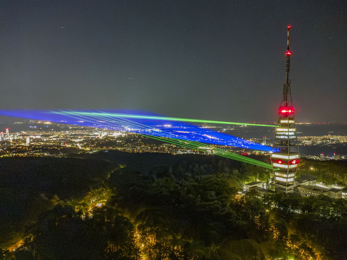 Blue and green beams cross from the tower over the city, the hillside forest lit gold beneath.