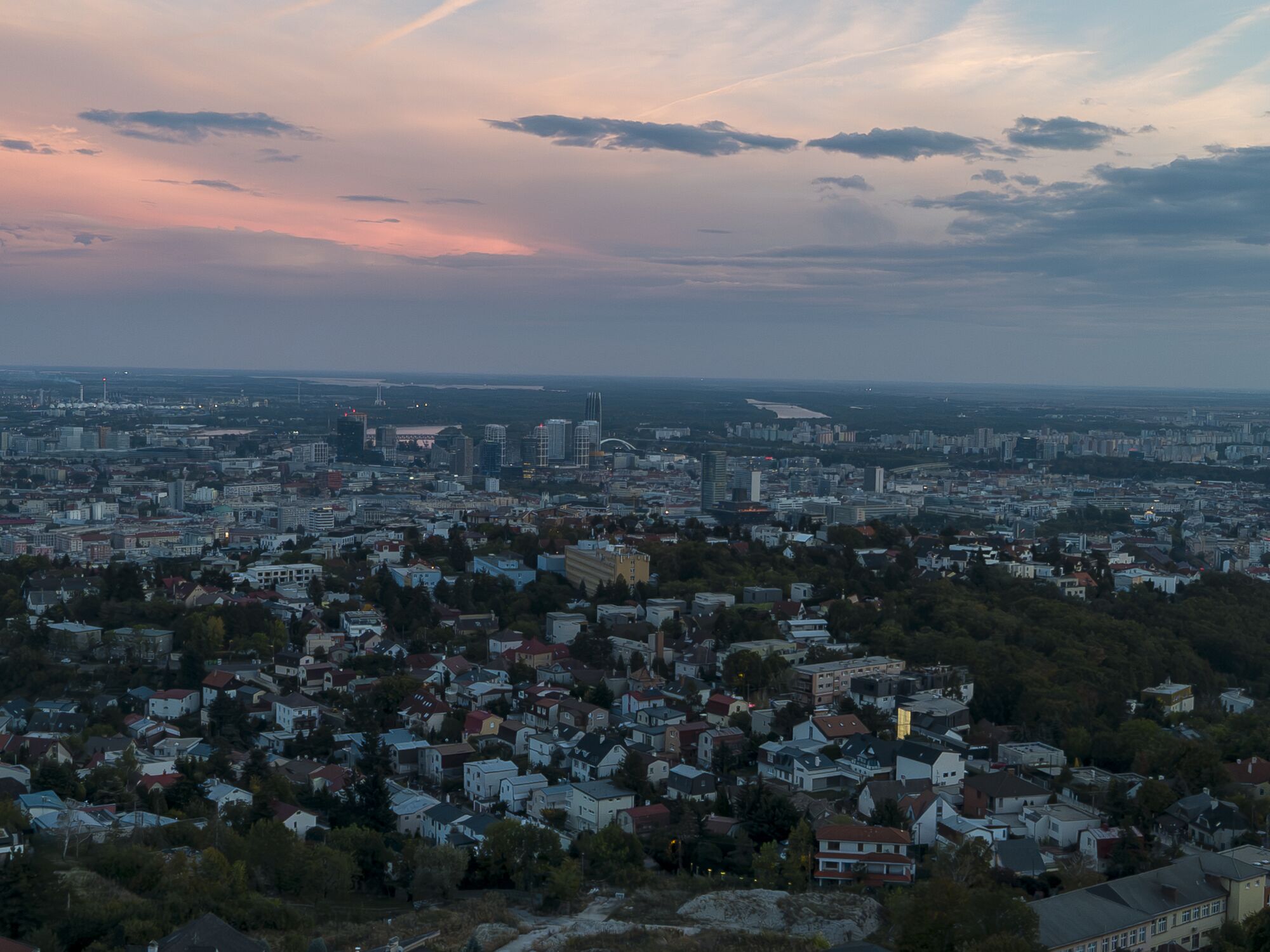 Bratislava from the air at dusk: red-roofed houses and the city beyond, under pink-lit streaks of cloud.